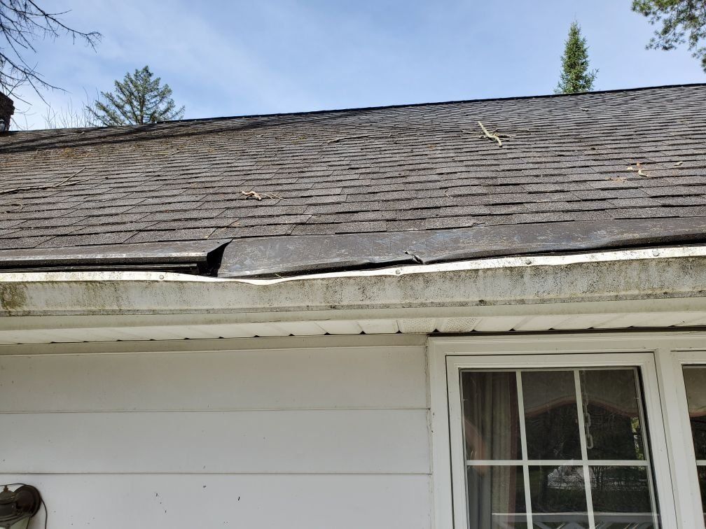 A white house with a roof damaged by a falling tree, missing shingles and a window.