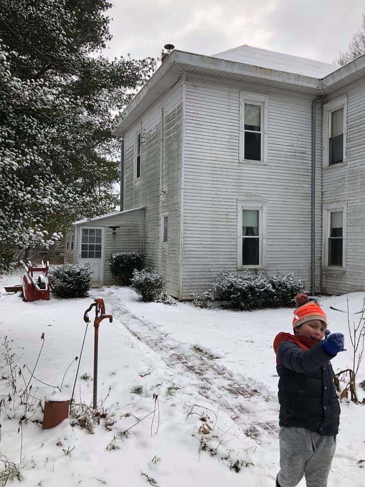 A young boy is standing in the snow in front of a white farmhouse.