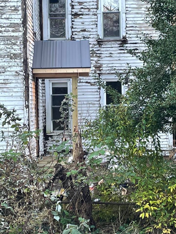 A white farmhouse with a metal roof is surrounded by trees and brush.