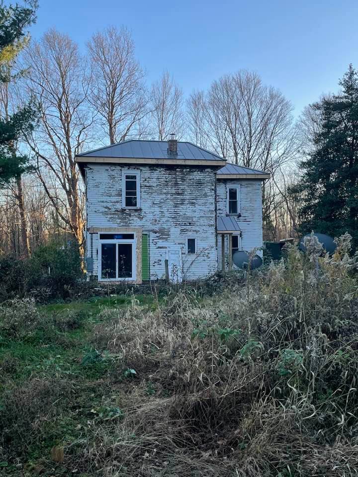 An old white farmhouse is sitting in the middle of a field surrounded by trees.
