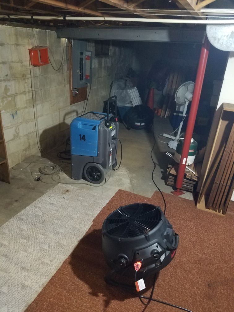 A fan is sitting in a basement next to an industrial machine and fan. 