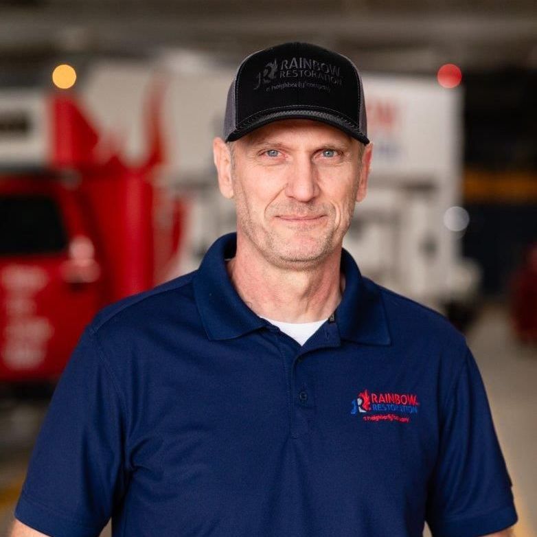 A man wearing a hat and a blue Rainbow Restoration shirt is standing in a parking garage and smiling.