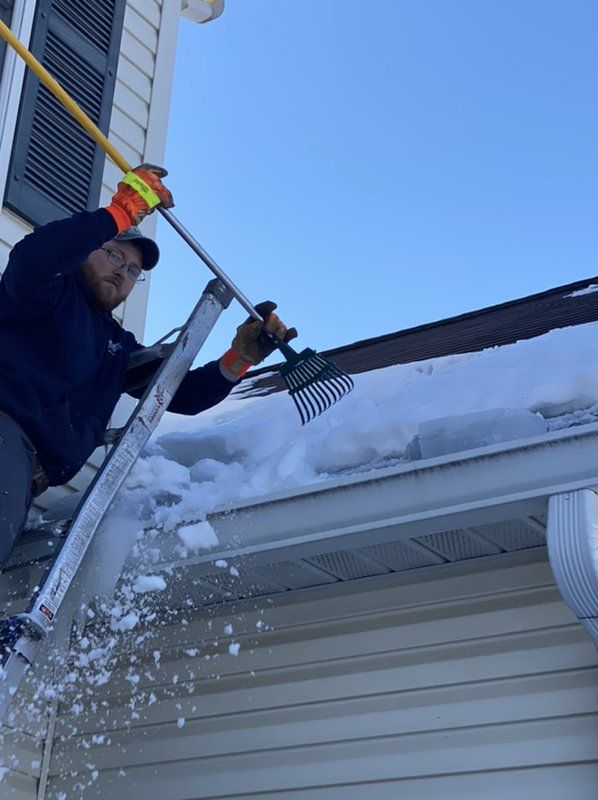 A man is standing on a ladder and raking snow off the roof of a house.