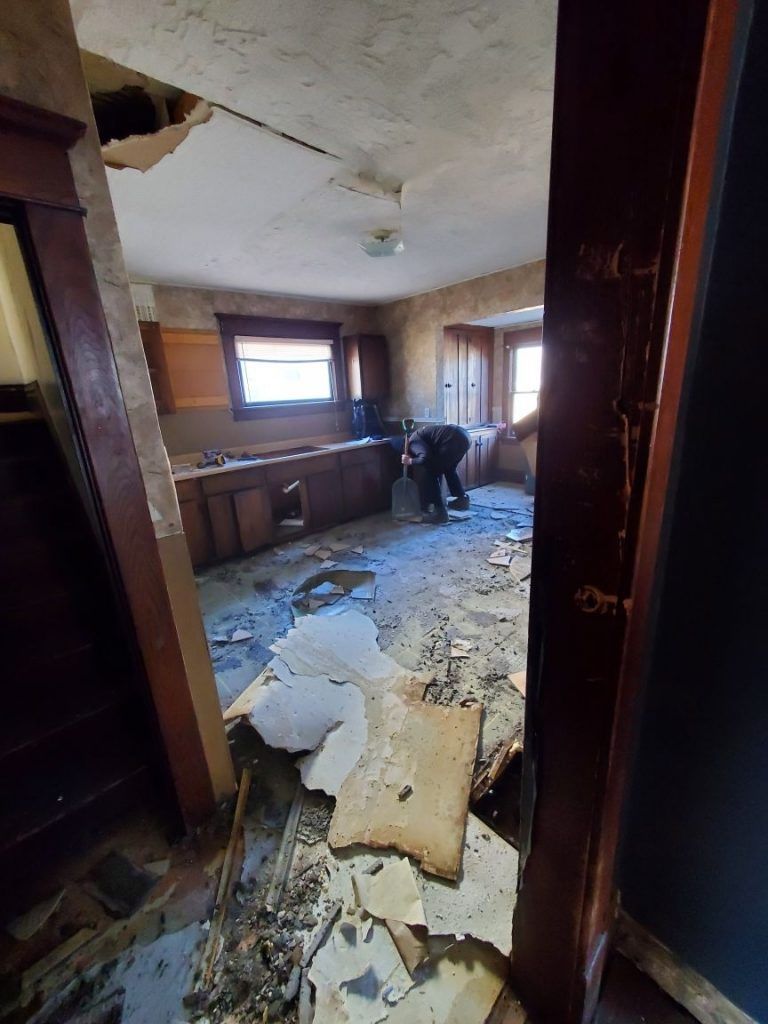 A man is kneeling in a messy kitchen with a hole in the ceiling.