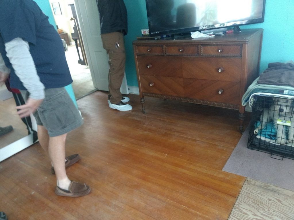 Two men are standing in a room with green walls, a wooden dresser, and a dog crate with white dog inside.