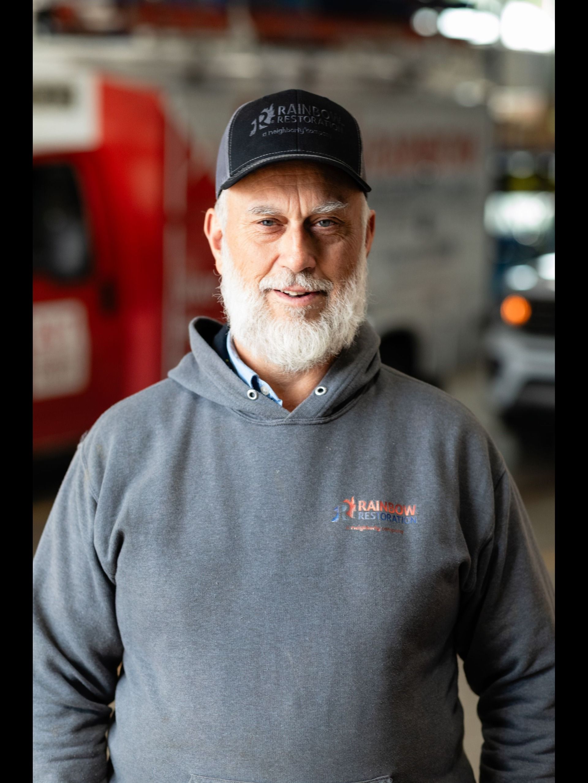 A man with a beard is wearing a hat and gray hoodie smiling for the camera.