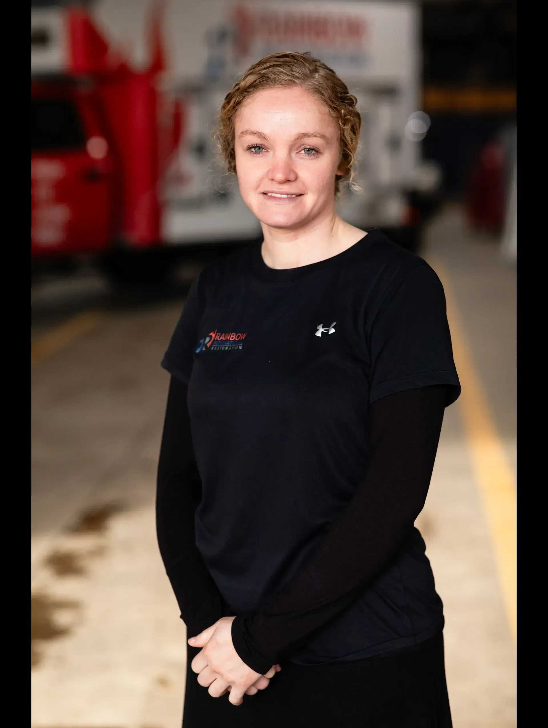 A woman wearing a black Rainbow Restoration shirt is standing in front of a truck smiling. 