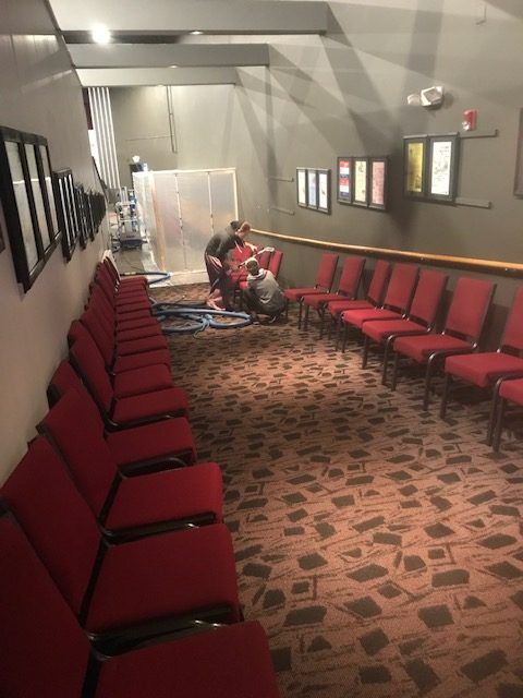 A row of red chairs are lined up in a room with a woman cleaning the wall.