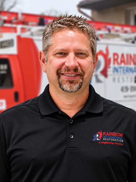 A smiling man with a beard is wearing a hat and a blue Rainbow Restoration shirt.