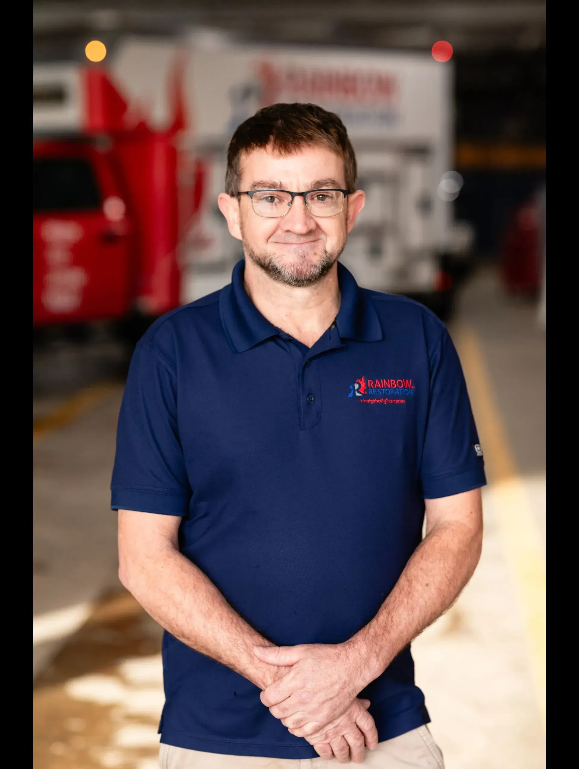 A man smiling wearing glasses and a blue shirt is standing in front of a red truck.