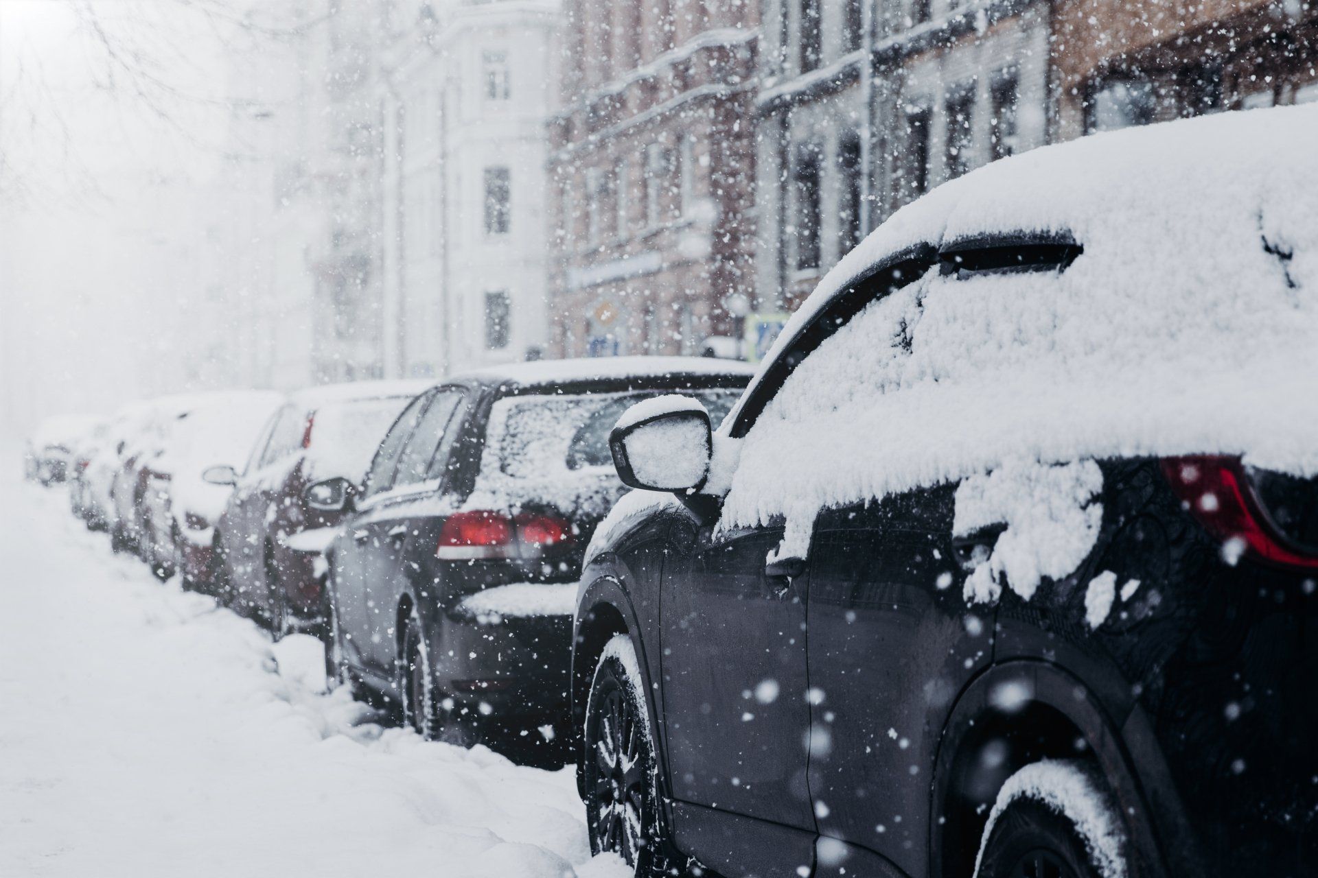 A row of cars are covered in snow on a snow covered street.