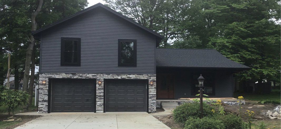 A black house with a concrete driveway and two garage doors is surrounded by trees.