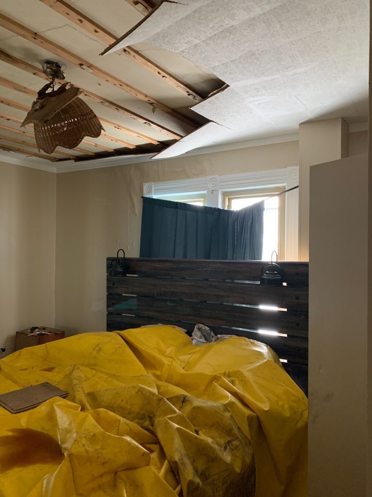 A room with a yellow tarp on the floor, a ceiling fan, and a water damaged ceiling. 