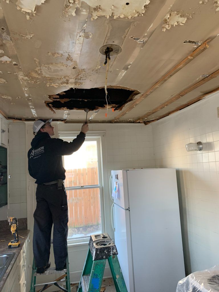 A man is standing on a ladder in a kitchen repairing a hole in the ceiling.