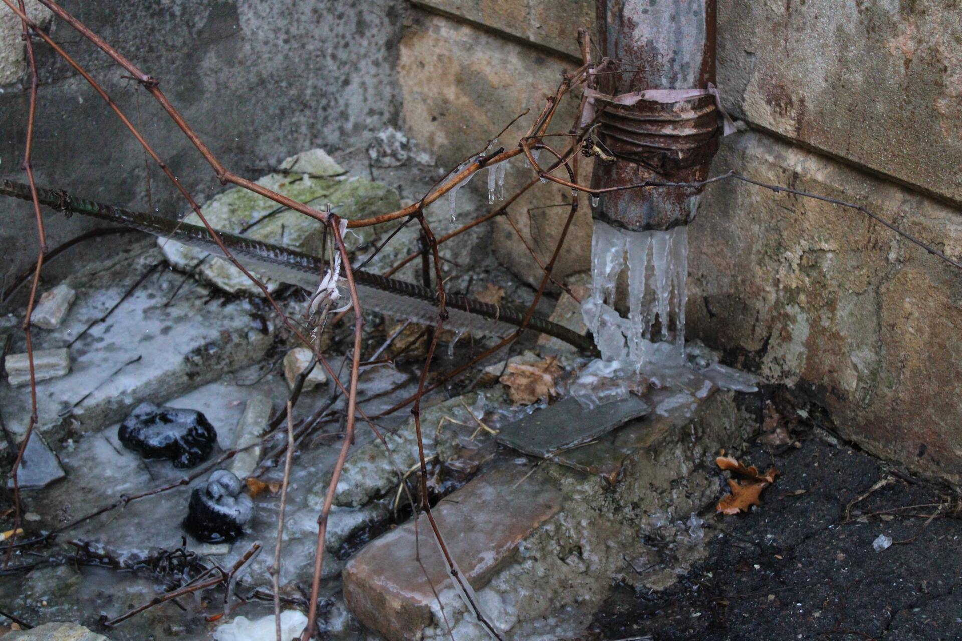 A frozen drainpipe with icicles coming out of it and a brick wall in the background.