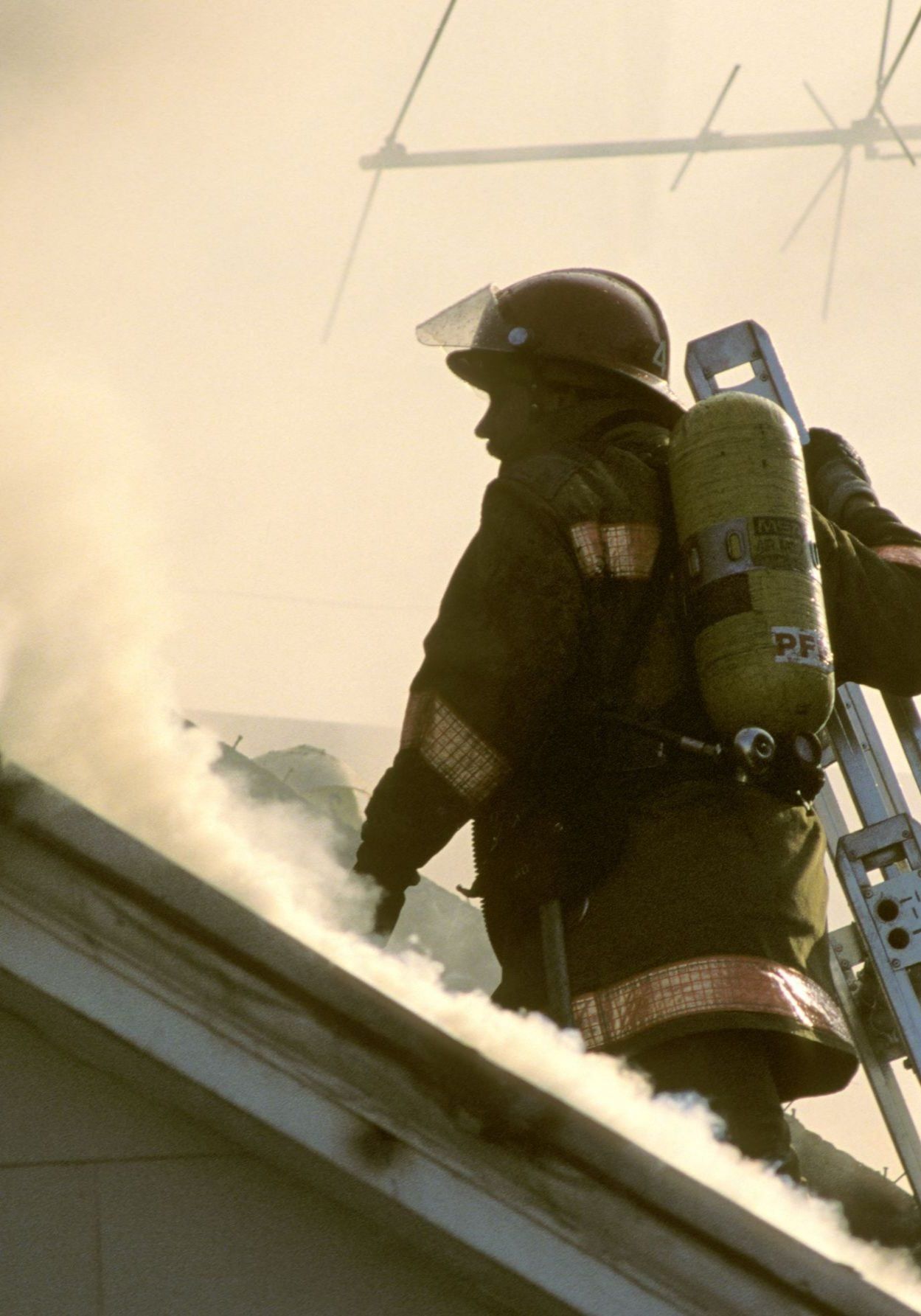 A fireman is on the roof of a building with smoke coming out of it.