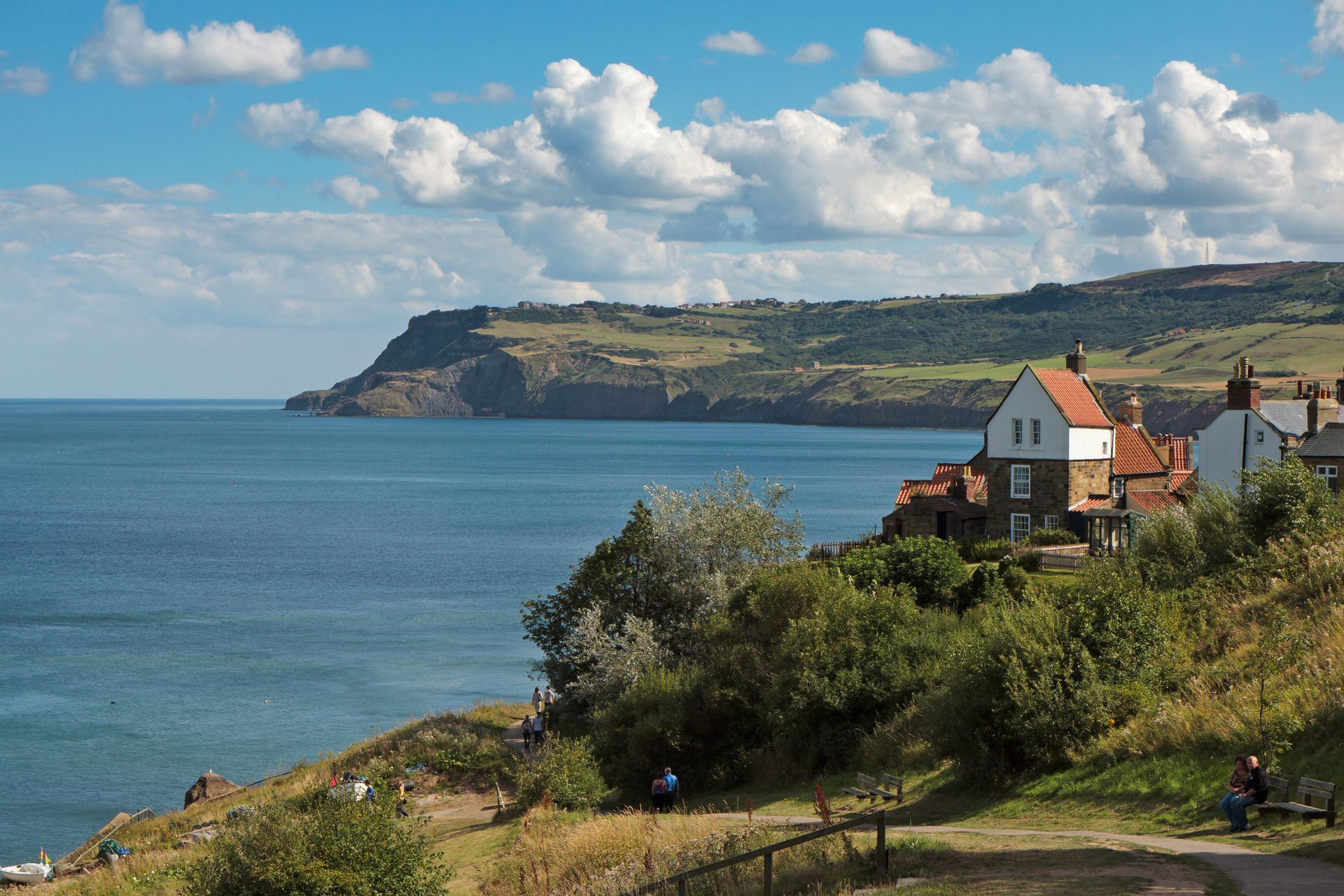 Coastal view: buildings on a cliffside overlooking a calm blue sea, with a distant cliff in the background, under a blue sky