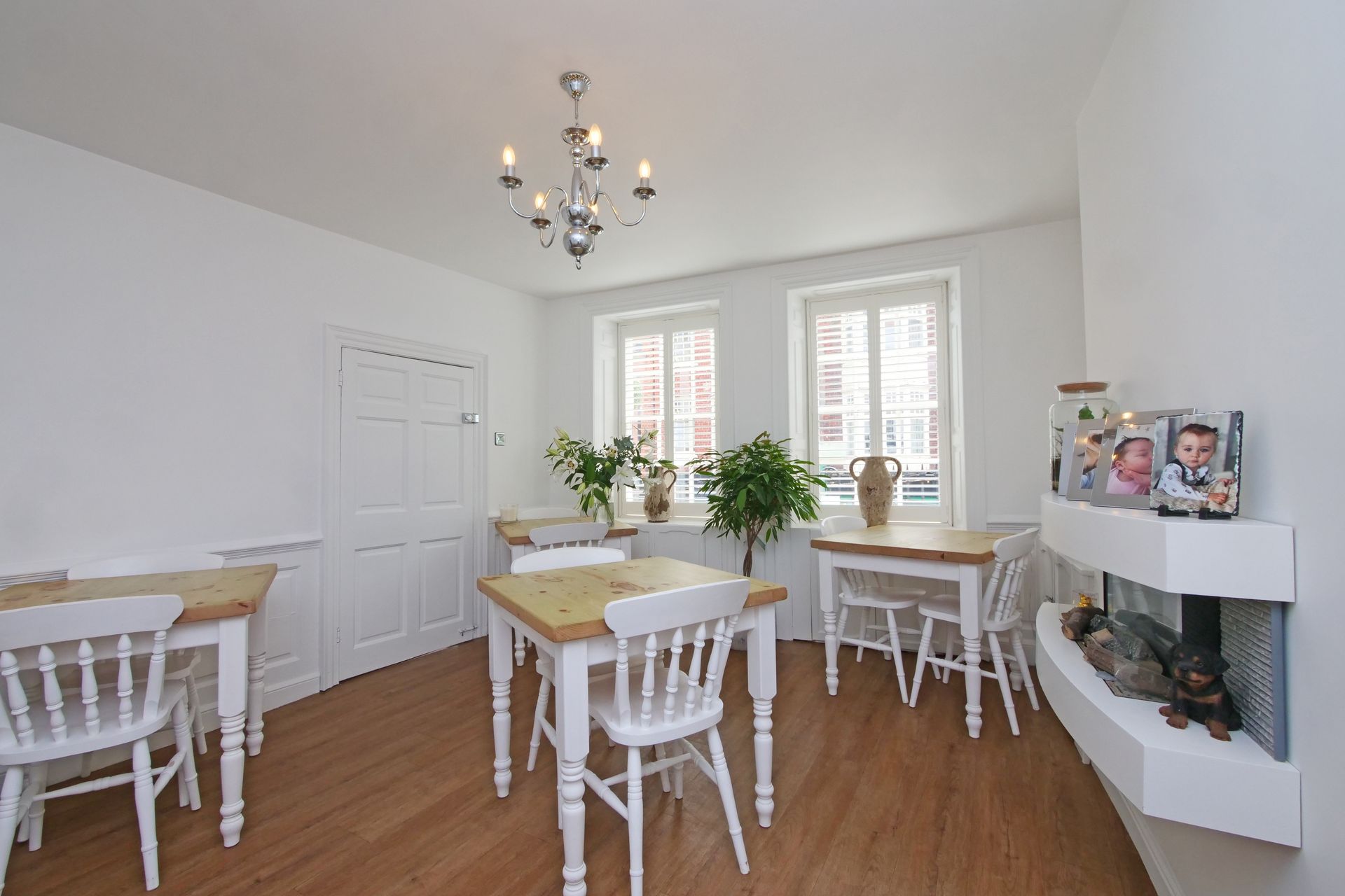 Bright dining room with white walls, tables, and chairs, a fireplace, and a chandelier. Two windows offer exterior views.