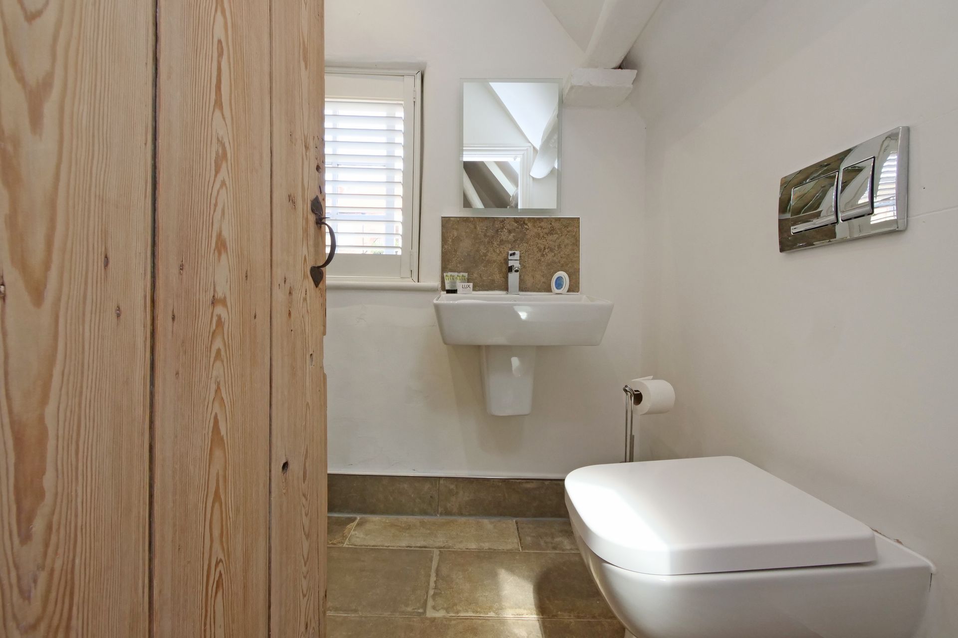 A small, bright bathroom with white walls, a white toilet, a sink, and a window with blinds; a wooden door is on the left.