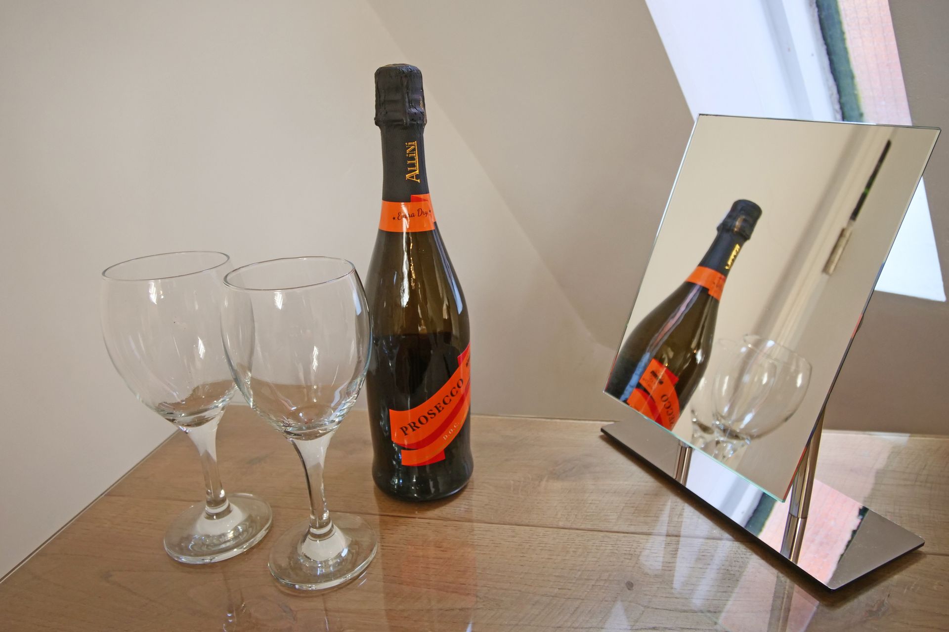 A bottle of Prosecco and two wine glasses sit on a wooden surface, with the bottle and a glass reflected in a nearby mirror.