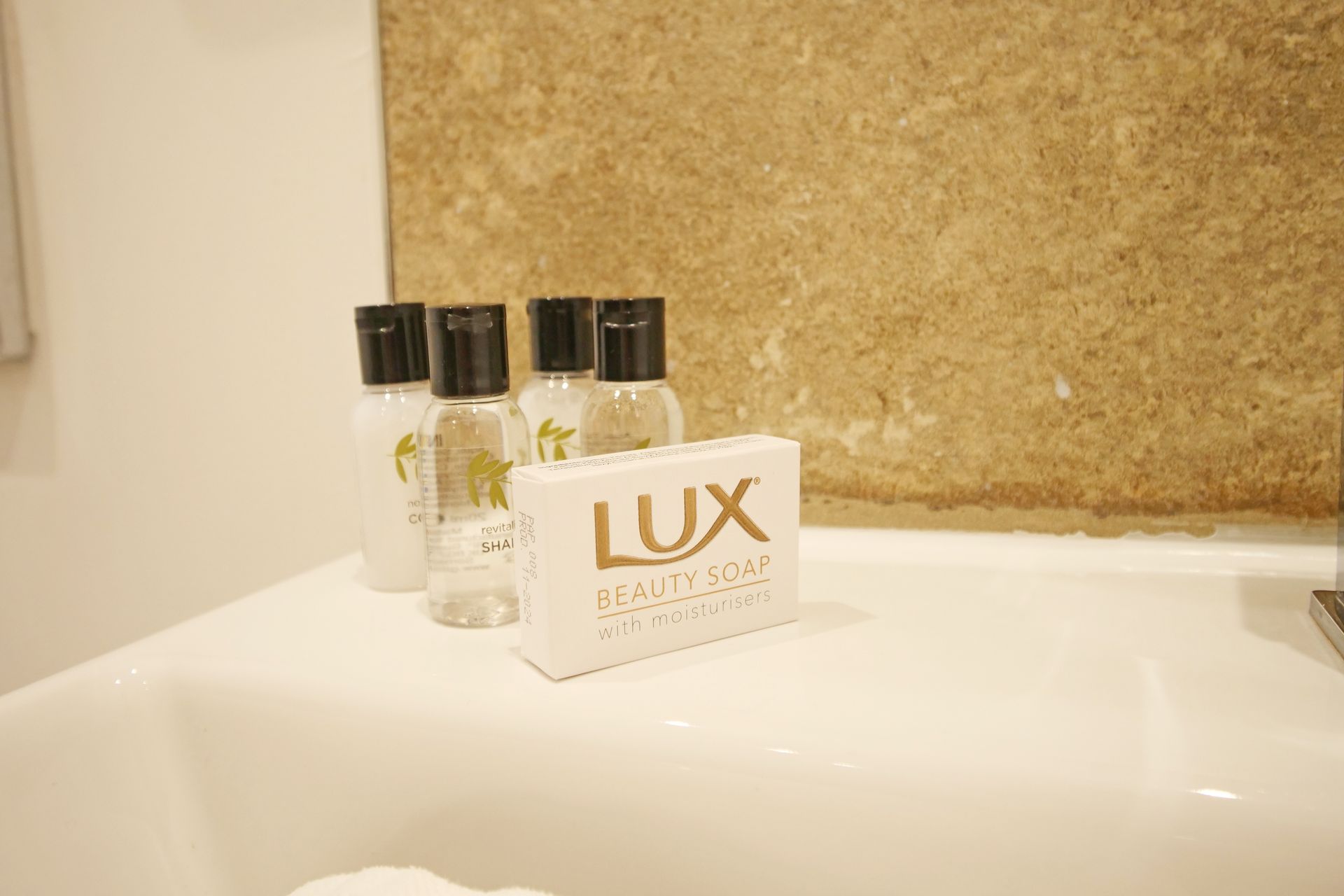 White sink with toiletries: small bottles of shampoo and a bar of Lux soap sit against a textured beige wall.