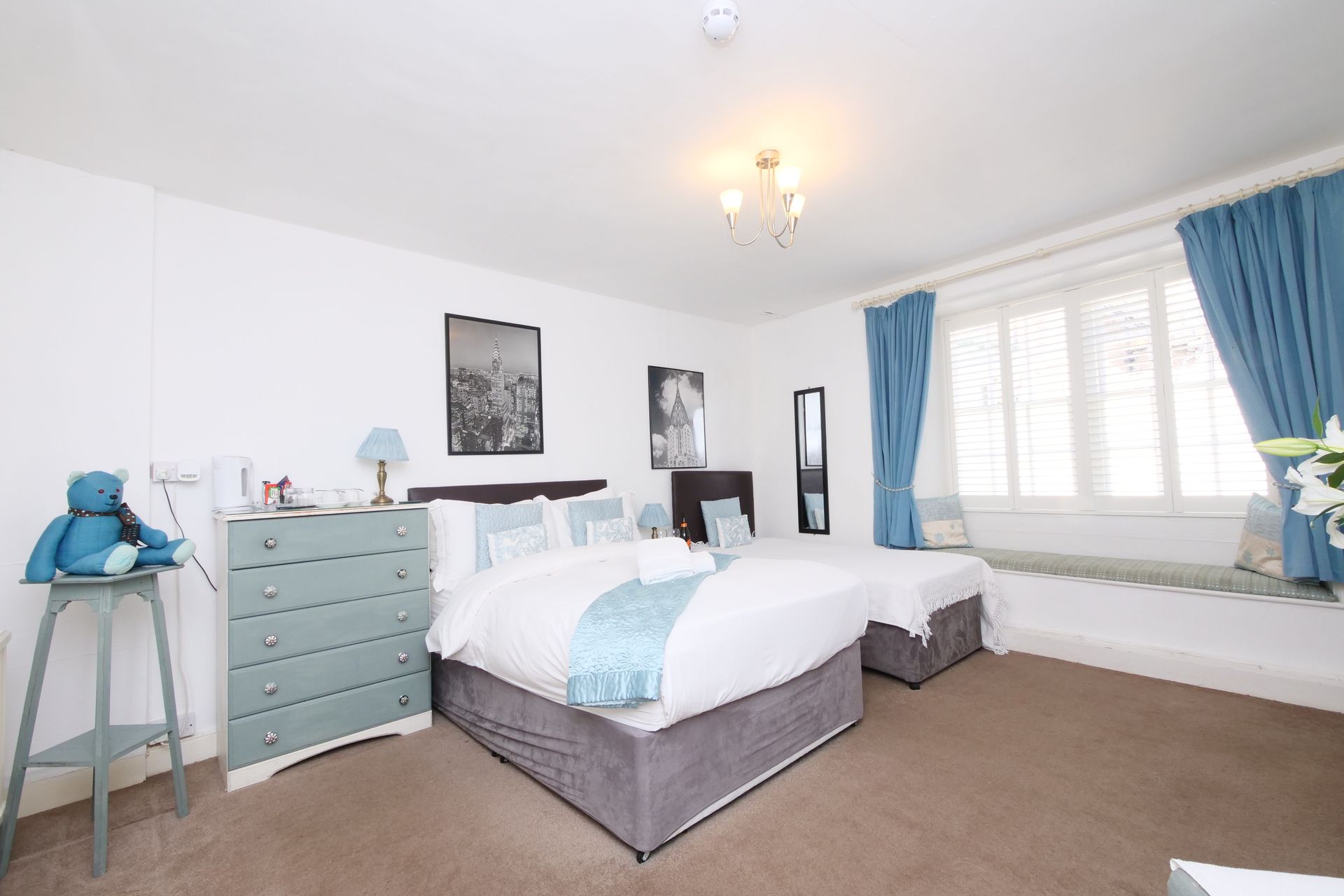 A bedroom with two beds, a dresser, and blue curtains. There is a teddy bear on a side table.