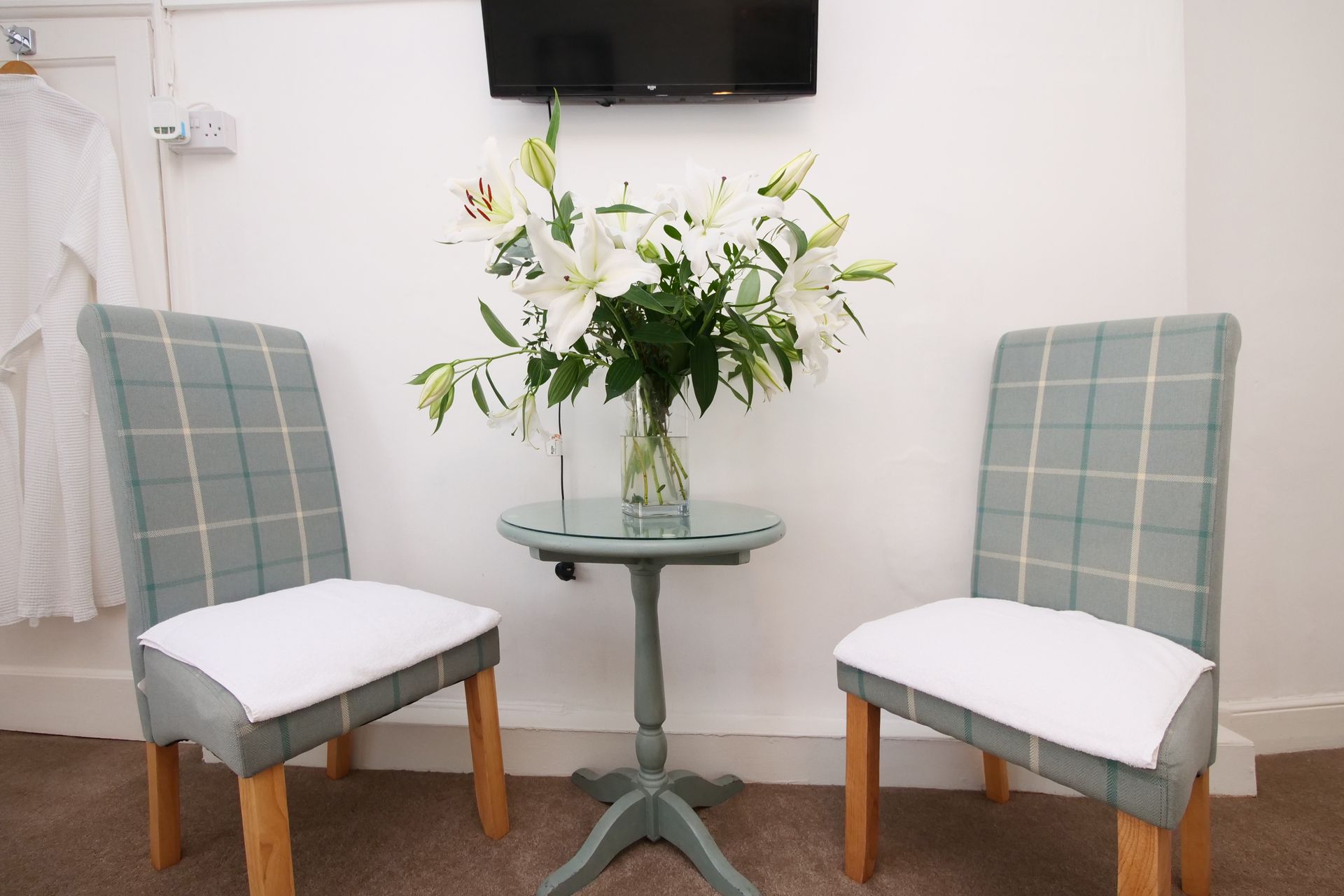 Two chairs flank a small table with a vase of lilies, beneath a wall-mounted TV. The chairs have light blue plaid upholstery.