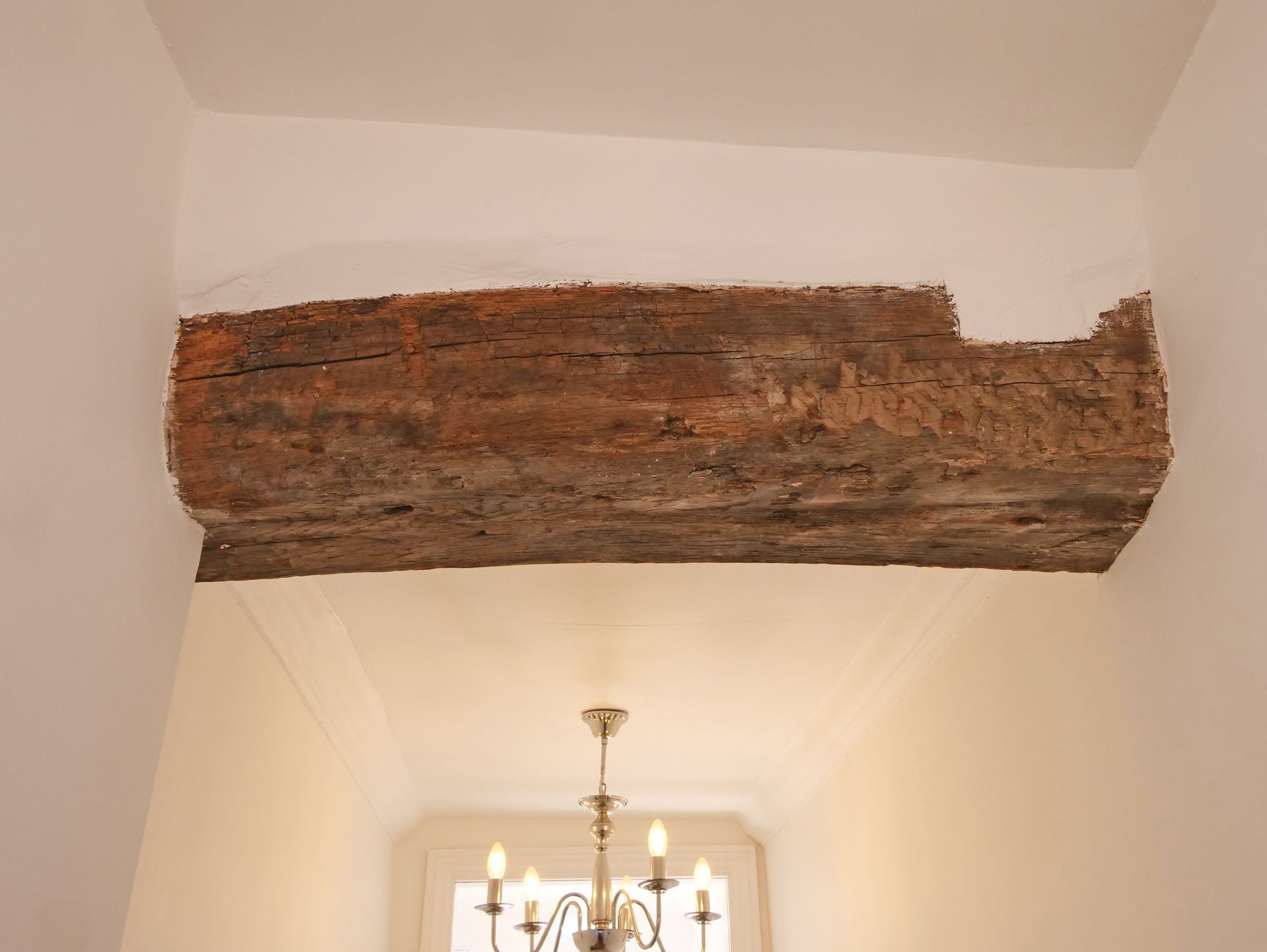 Wooden beam in white-painted room, positioned above a hallway with a chandelier.
