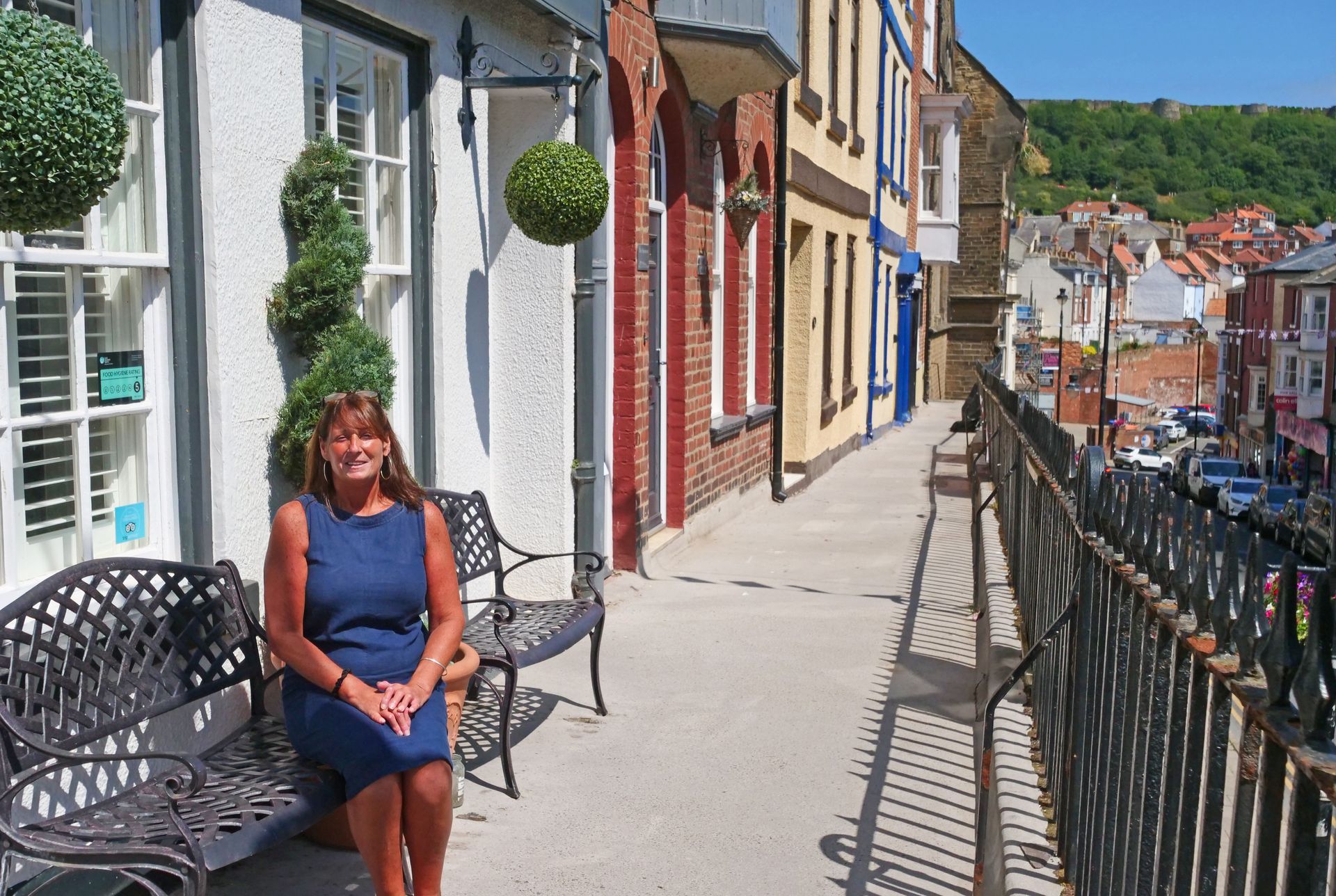 Woman in blue dress sits on a bench on a sunny street, overlooking a town. Buildings in the background.