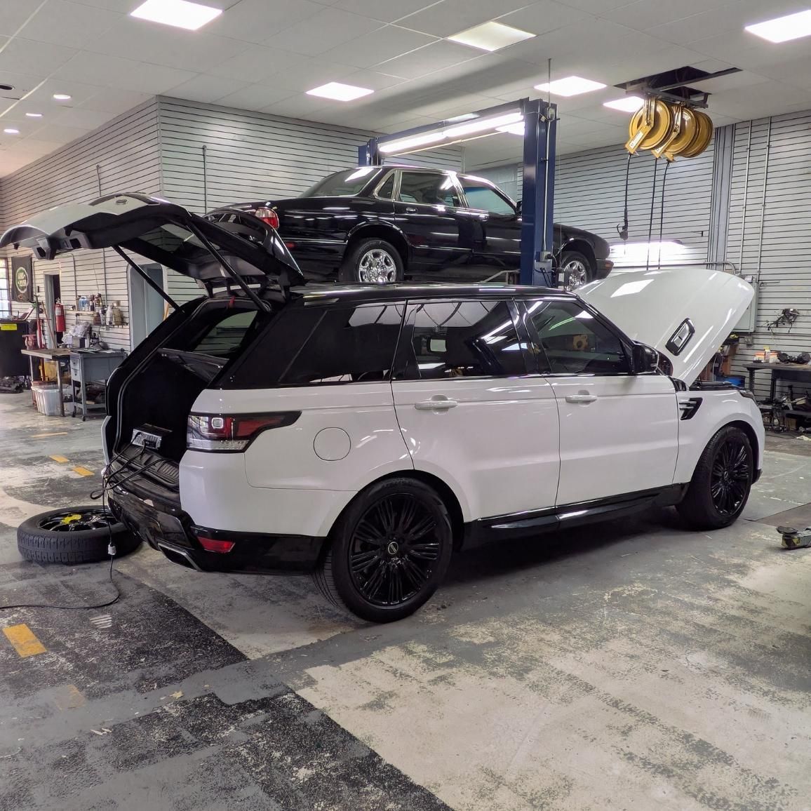 White SUV with doors and hatch open in an auto repair shop, with another car on a lift behind it.