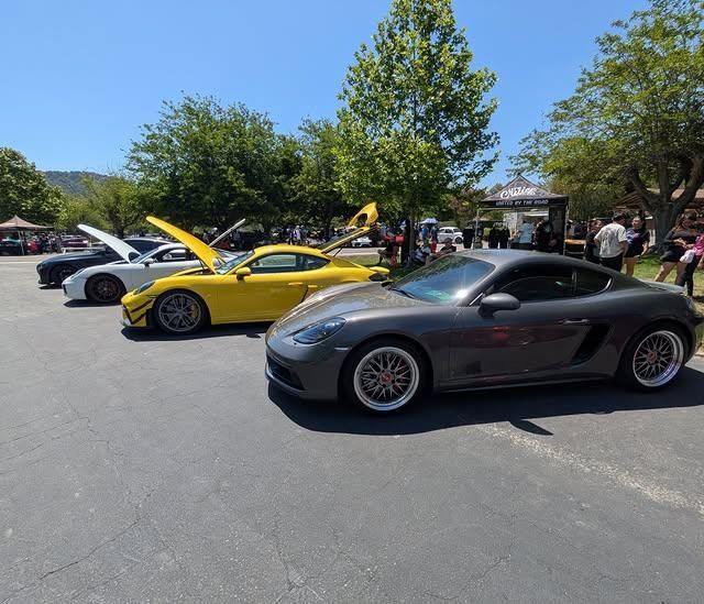 Two sports cars at an outdoor car show, a black coupe in front of a yellow car with its hood open.
