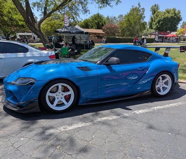 Blue sports coupe parked outdoors on a sunny street, with white wheels and trees in the background