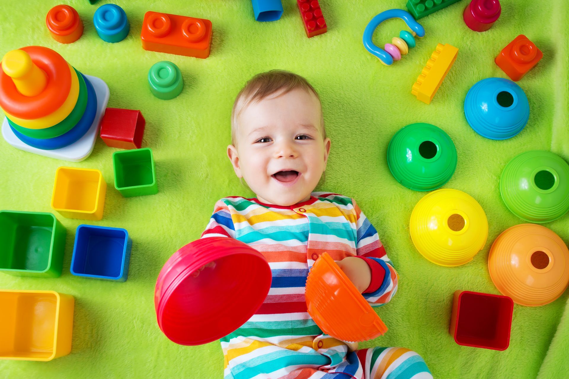 Smiling baby lying on a green mat, surrounded by colorful toy blocks and rings.