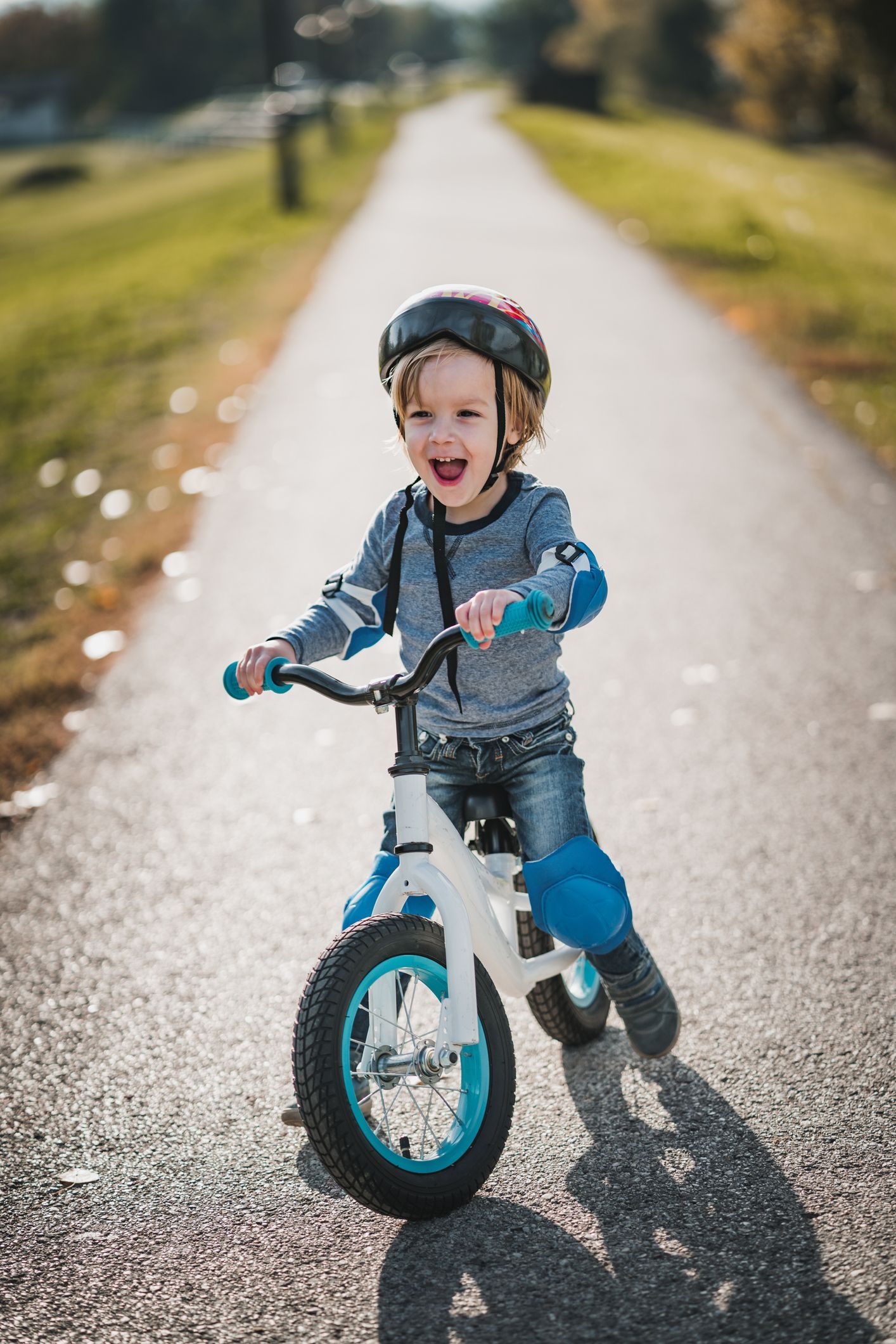 Child riding a blue balance bike on a sunlit path, smiling toward the camera.