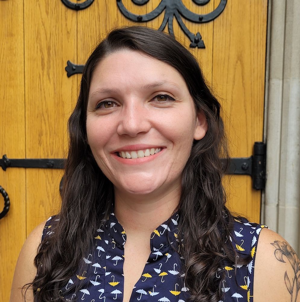 Woman smiling, dark hair, patterned top, standing in front of a wooden door with decorative hardware.