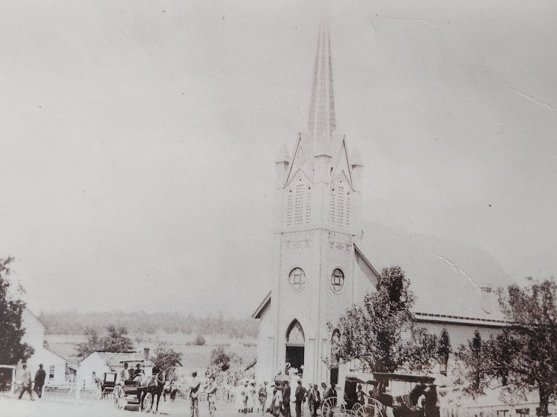 A tall church with a steeple; people gather outside. Horse-drawn carriages line the road.