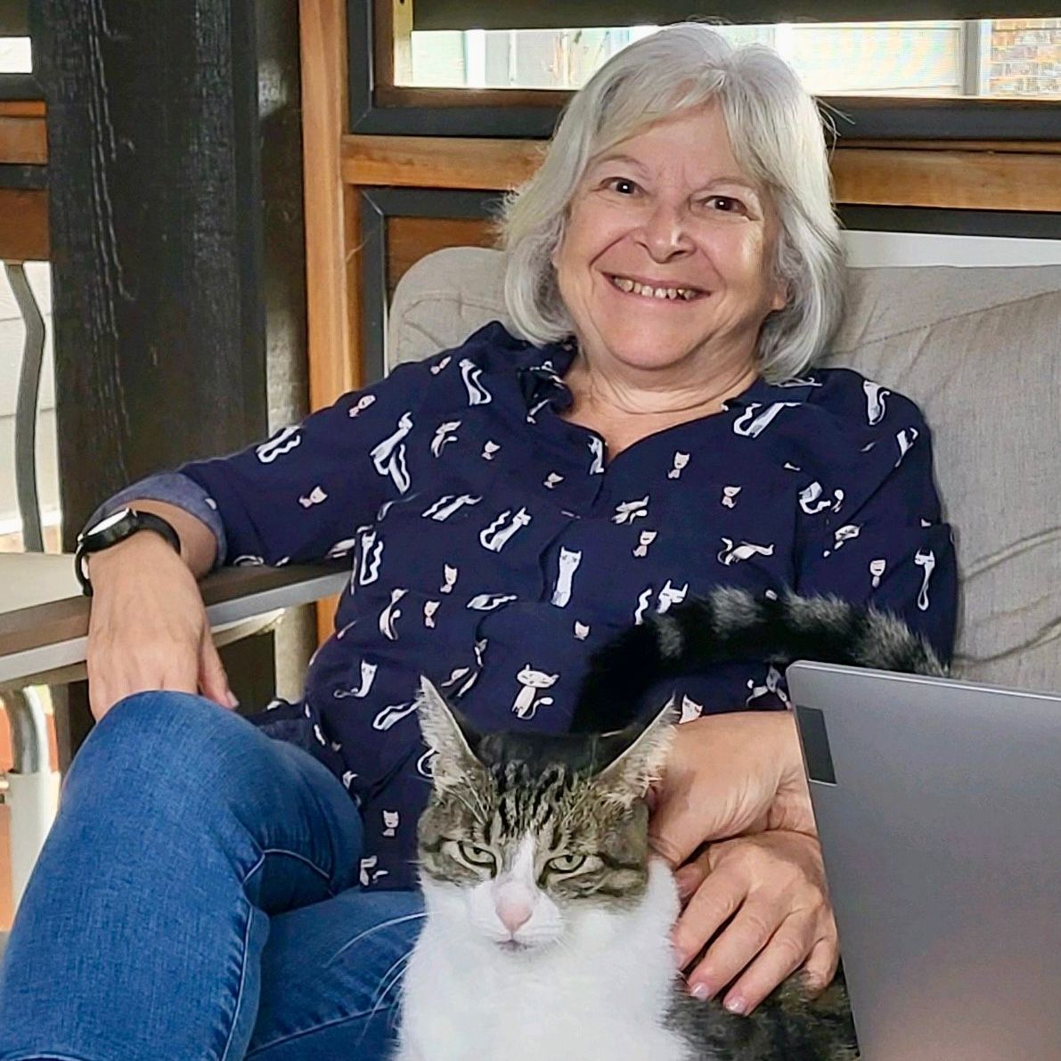 Woman with gray hair and cat on her lap, smiling, wearing a blue shirt, sitting outside.