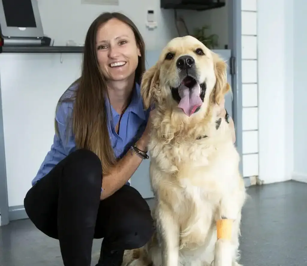 Woman kneeling next to a golden retriever; both smiling. Dog has a bandaged leg in a veterinary setting.