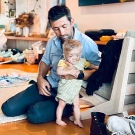 Man kneeling, holding a toddler with a medical tube. The child is wearing a green onesie. Indoors, warm tones.
