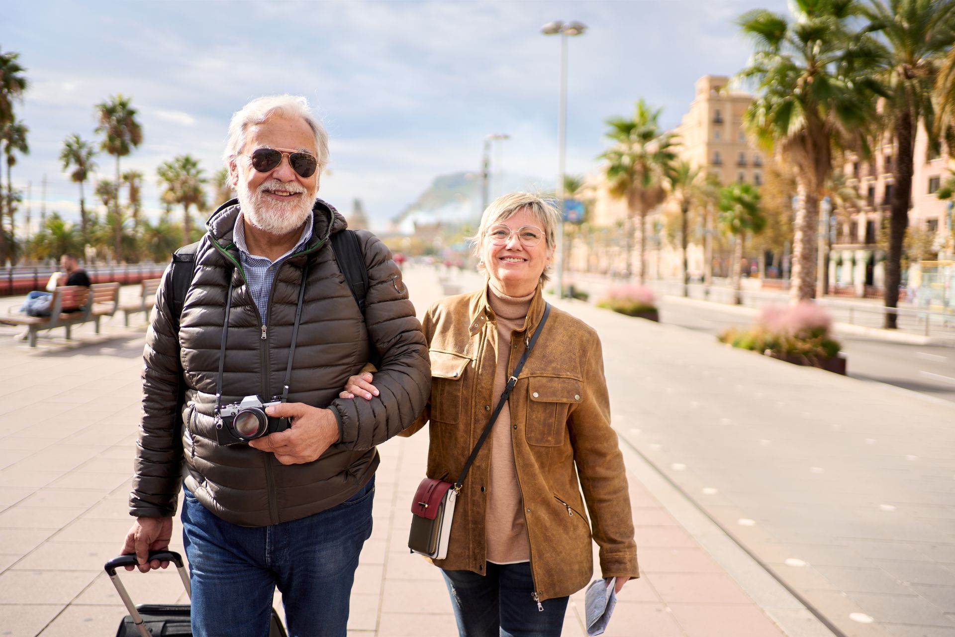An older couple walks arm-in-arm along a palm-lined promenade, carrying luggage and a camera on a sunny day.