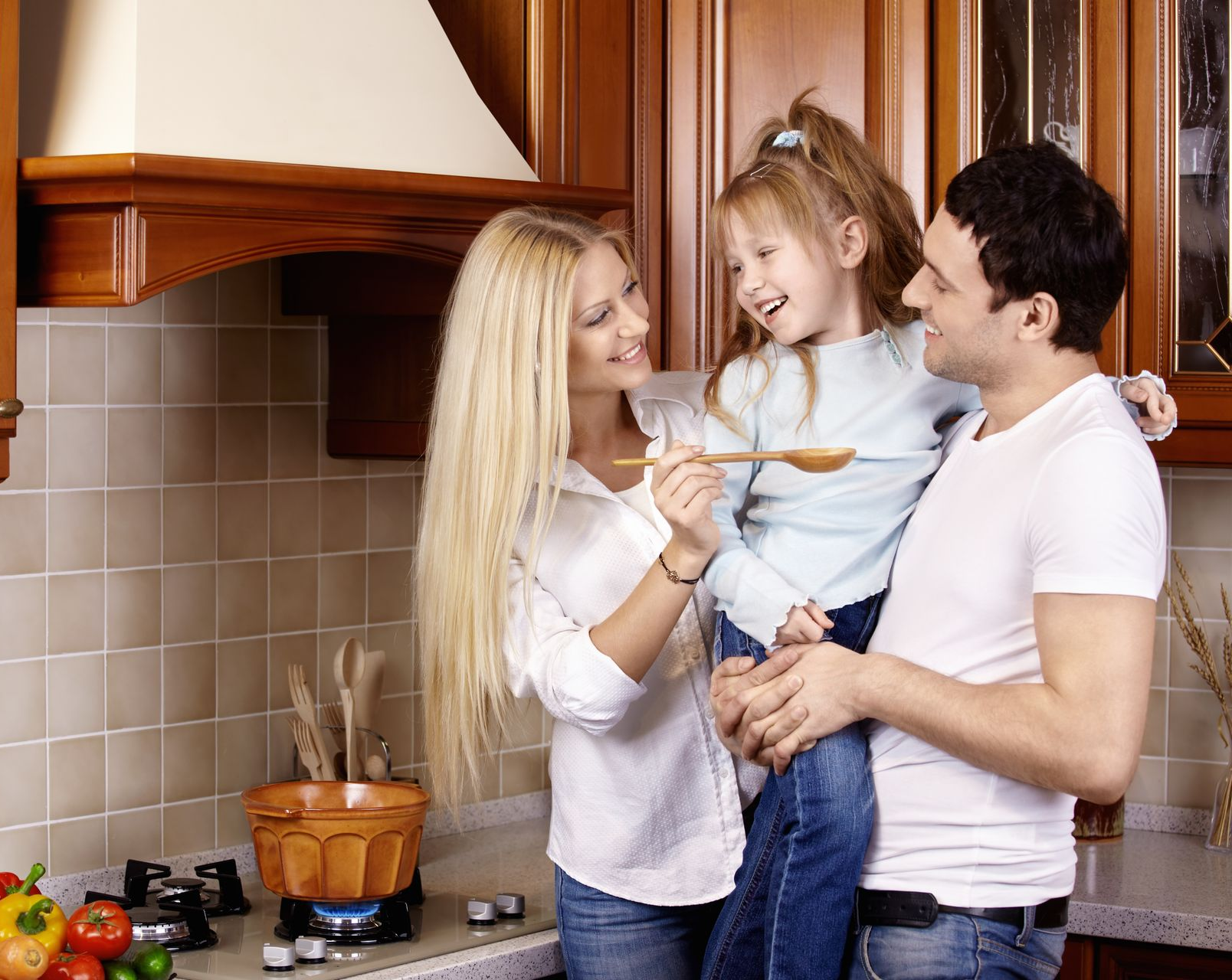 A family standing in a kitchen, with a person holding a wooden spoon to a child's mouth while they all smile.