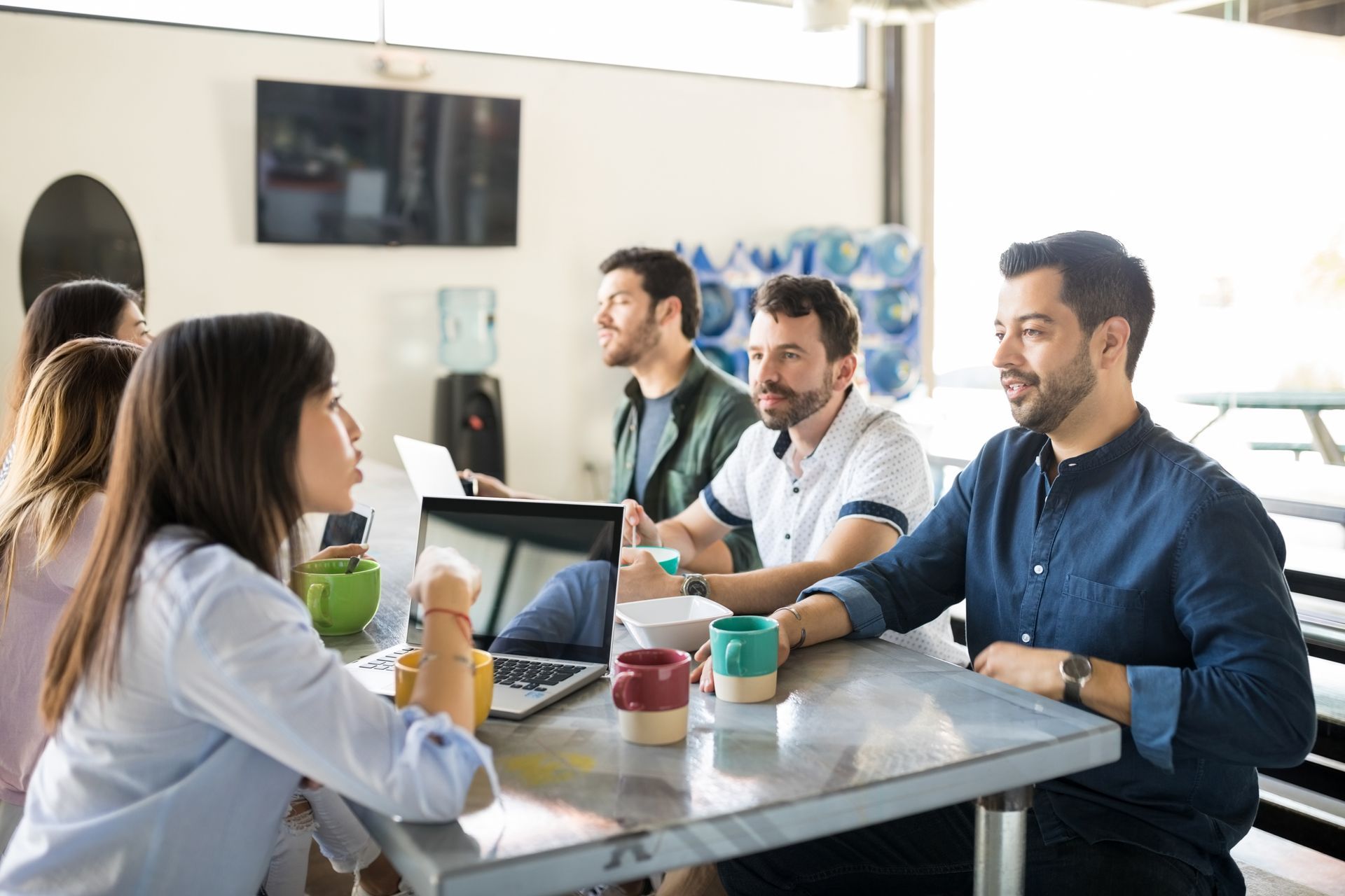 A group of people sit around a table in a modern, well-lit office, collaborating and talking while using a laptop.