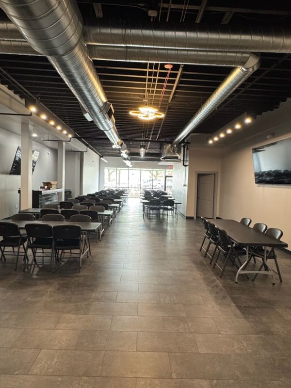Long banquet hall with rows of black folding tables and chairs under exposed ducts and ceiling lights