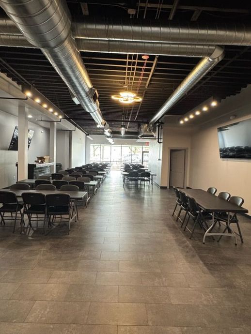 Long banquet hall with rows of black folding tables and chairs under exposed ducts and ceiling lights
