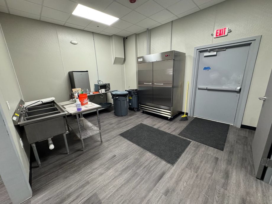 Commercial kitchen area with stainless steel carts, prep table, trash can, and gray door under exit sign.