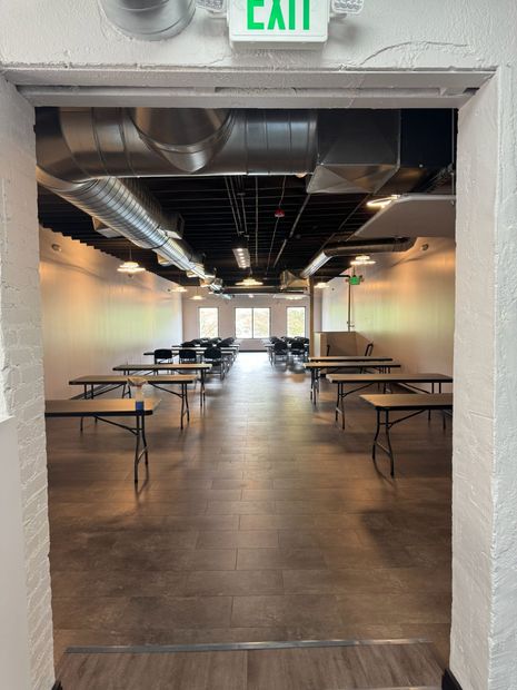Empty event space with folding tables, exposed ceiling, and bright windows at the far end.