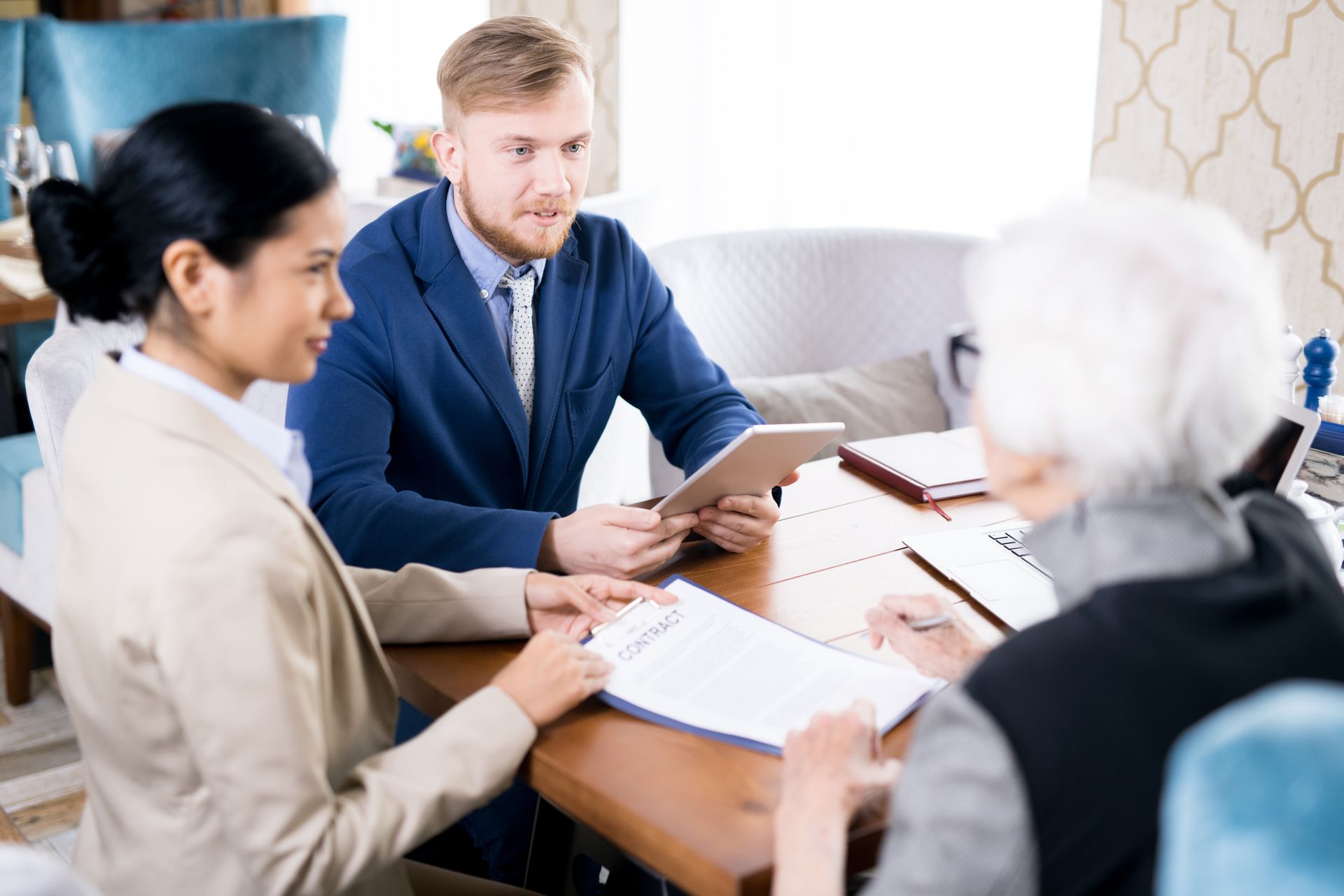 Three people at a table reviewing documents. Man in suit holds tablet; woman on the left reviews papers. An older person signs.