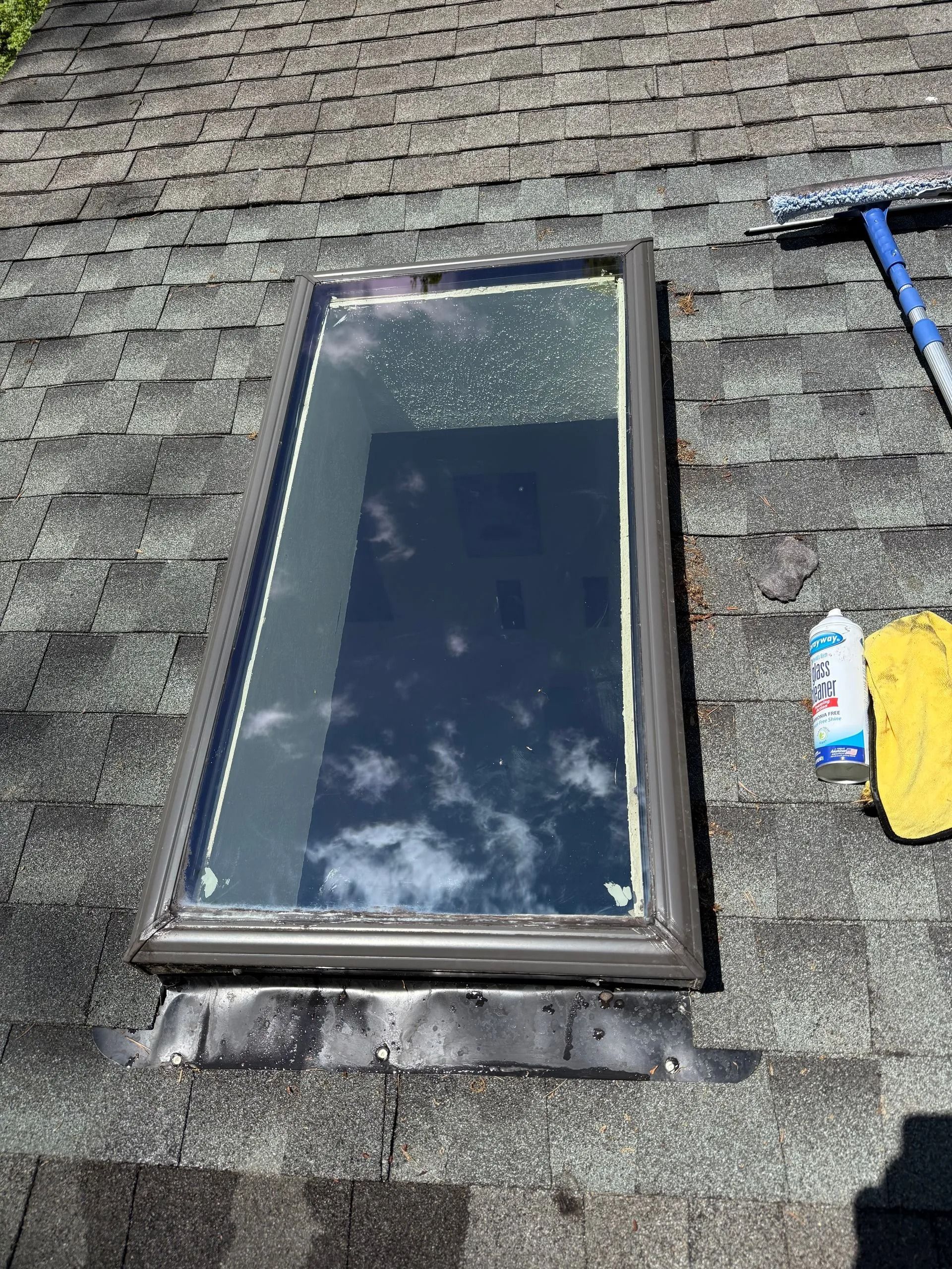 A skylight on an asphalt shingle roof with a squeegee, cleaning spray, and cloth nearby.