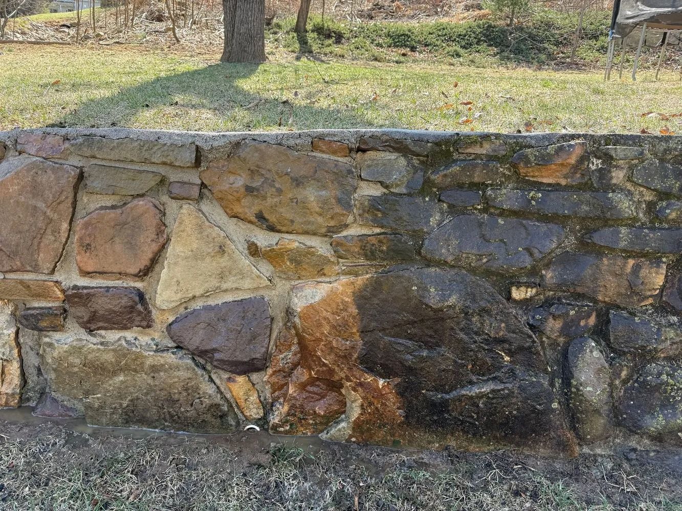 A textured stone retaining wall with various sizes of brown and gray rocks set in mortar, situated before a grassy lawn.