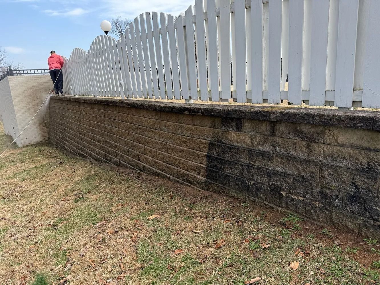 A person in a red jacket pressure washes the dark, stained sections of a concrete retaining wall under a white fence.